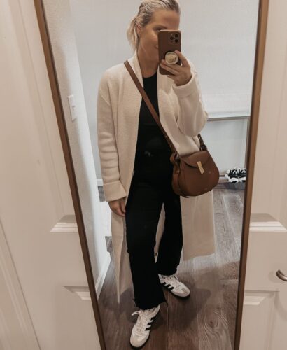 Lindsey Denver wearing a white coatigan, paired with a black turtleneck sweater, dark wash jeans, and black casual sneakers. She accessorizes with a brown leather tote bag and gold hoop earrings.