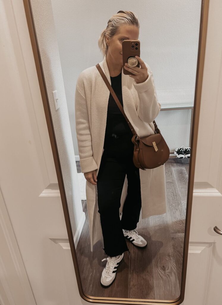 Lindsey Denver wearing a white coatigan, paired with a black turtleneck sweater, dark wash jeans, and black casual sneakers. She accessorizes with a brown leather tote bag and gold hoop earrings.