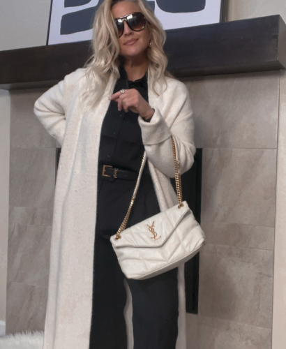 "Image of Lindsey Denver wearing a black monochromatic outfit with a white coatigan and carrying a YSL LouLou bag. She is standing outdoors against a brick wall with a confident expression on her face.