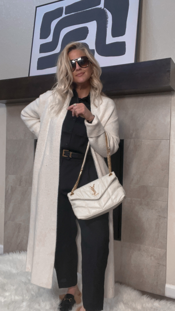 "Image of Lindsey Denver wearing a black monochromatic outfit with a white coatigan and carrying a YSL LouLou bag. She is standing outdoors against a brick wall with a confident expression on her face.