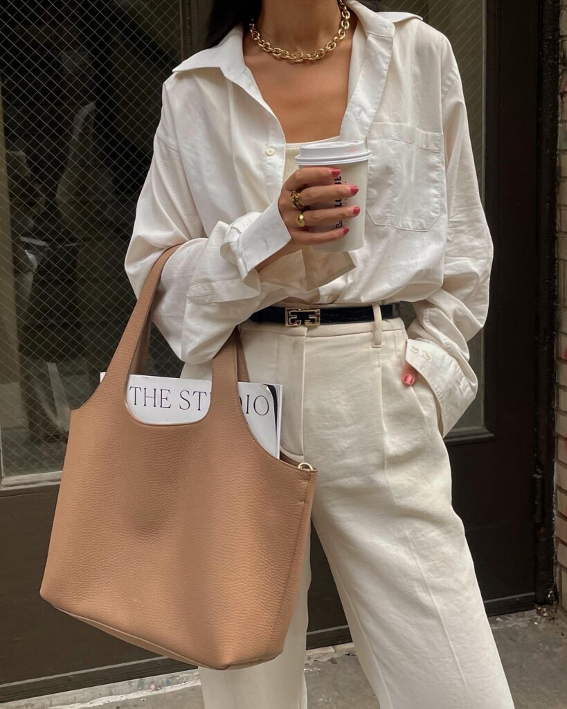 image of woman wearing sustainable clothing from Cuyana. Leather tote, white trousers and white linen top.